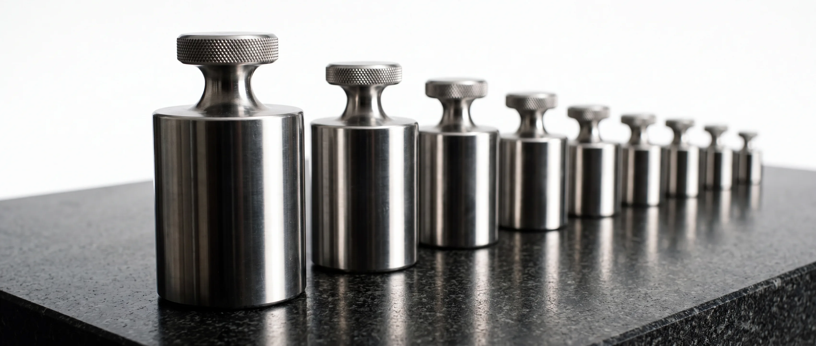 Nine polished steel calibration weights standing in a line on a black granite surface plate, each one smaller than the last, receding toward a white studio backdrop.